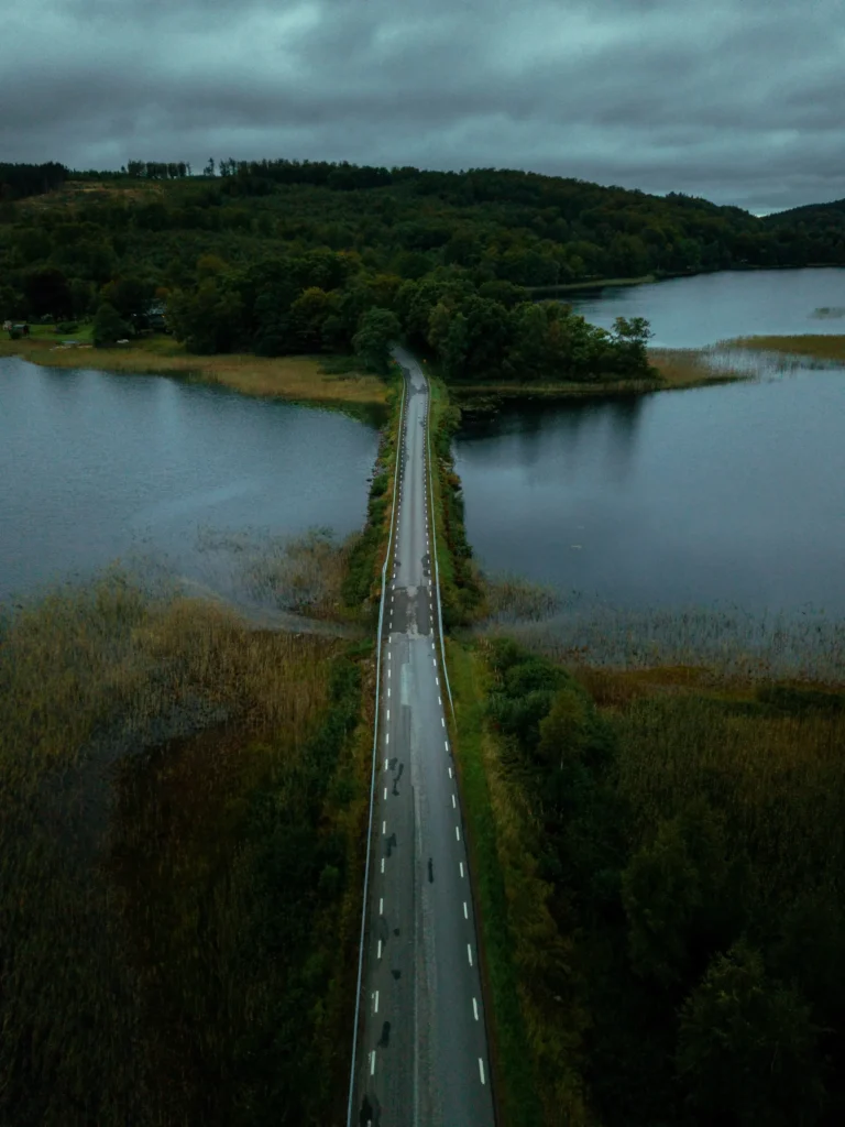 Ensam väg som sträcker sig över en spegelblank sjö i svensk landsbygd på en tidig höstmorgon, omgiven av träd i varma höstfärger under en molntäckt himmel.