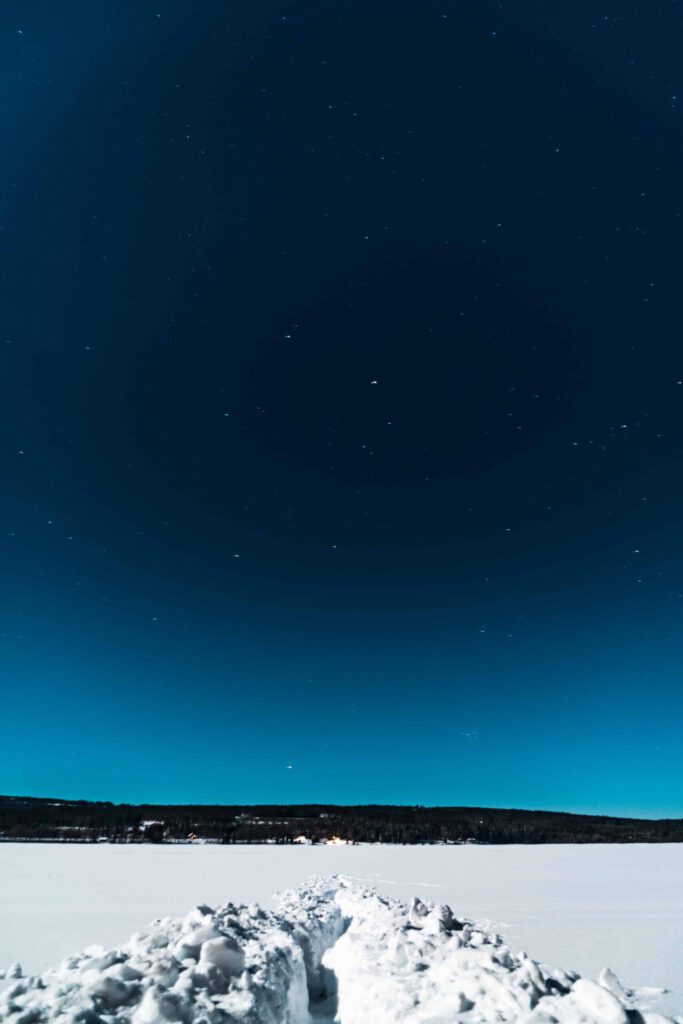 Stjärnklar vinternatt med ett snötäckt landskap och ett upptrampat spår som leder mot horisonten, där skog och små ljus från hus syns i fjärran.