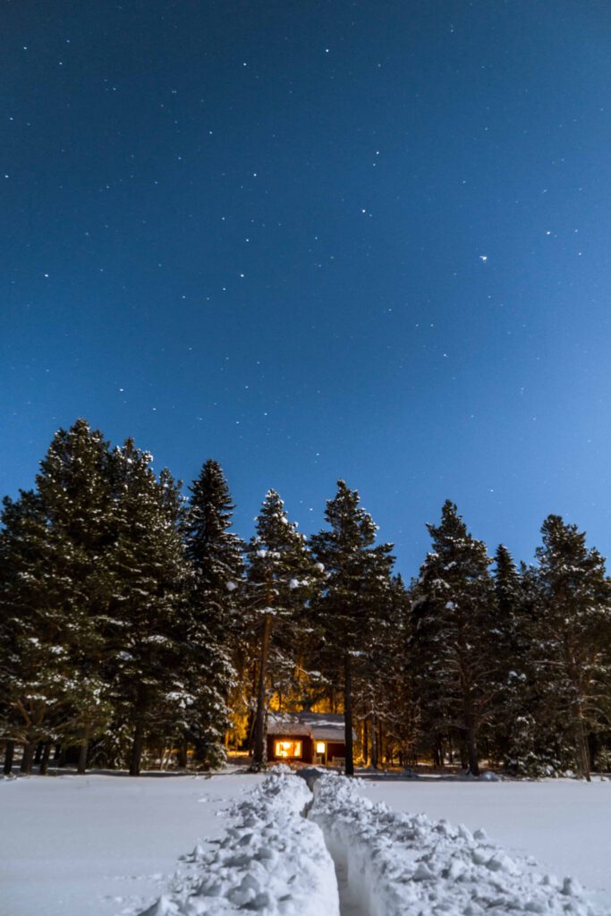 Stuga i en vinterskog med snötäckta träd och stjärnklar himmel, med ett upptrampat snöspår som leder fram till stugan vars fönster lyser varmt i natten.