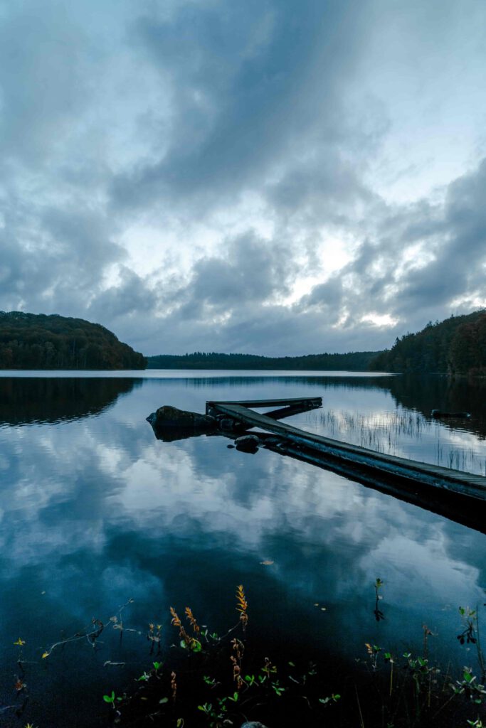 En lugn sjö i gryningens blå ljus, med en brygga som sträcker sig ut över det stilla vattnet. Himlen är molnig och atmosfären är kall och fridfull precis innan soluppgången.