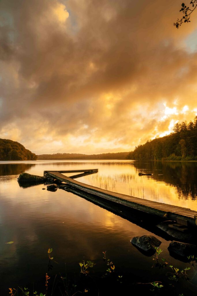 Poster som visar En brygga leder ut över en stilla sjö under en dramatisk soluppgång. Himlen exploderar i färger precis innan solen stiger över horisonten, med reflektioner i vattnet.