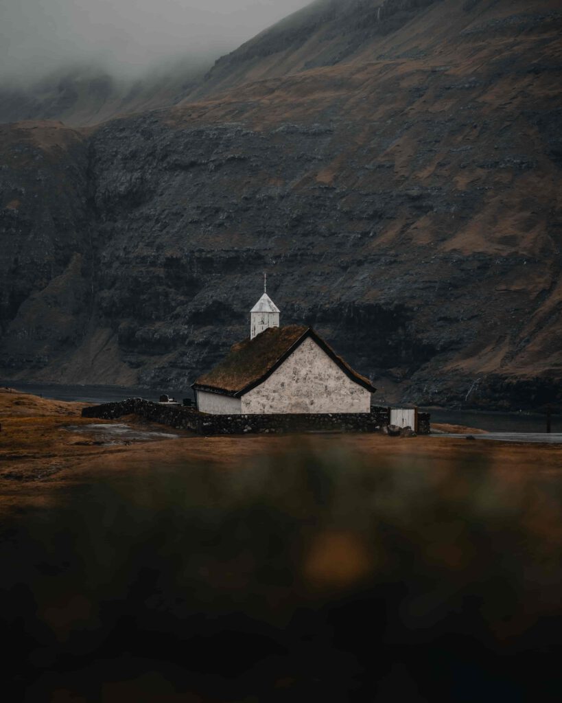 En vit, mossklädd stenkyrka står ensam mot en massiv, mörk bergsvägg i ett disigt och avlägset landskap.