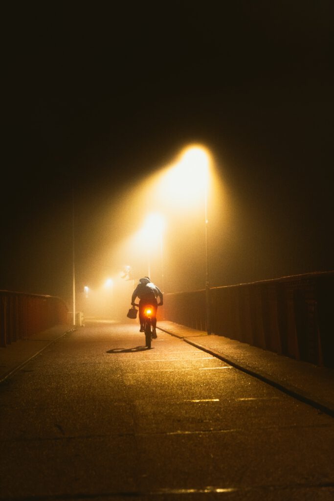 En cyklist på en dimmig väg under nattens mörker, upplyst av varma gatlampor och med ett rött bakljus som lyser svagt i natten.