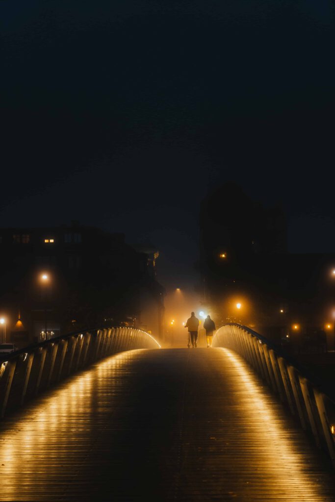 Två personer promenerar över en upplyst bro i en dimhöljd stadsmiljö på natten.