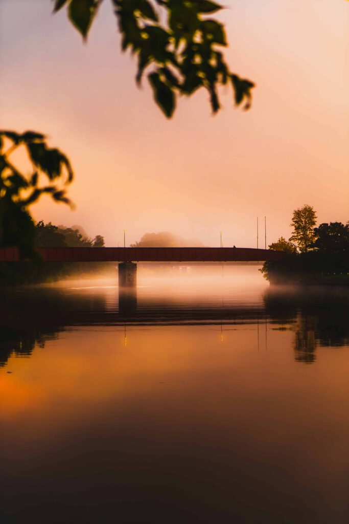 En dimmig bro över Nissan i Halmstad i gryningen med en varm, orange himmel som reflekteras i det lugna vattnet. Träd och löv ramas in bilden, vilket skapar en fridfull atmosfär.