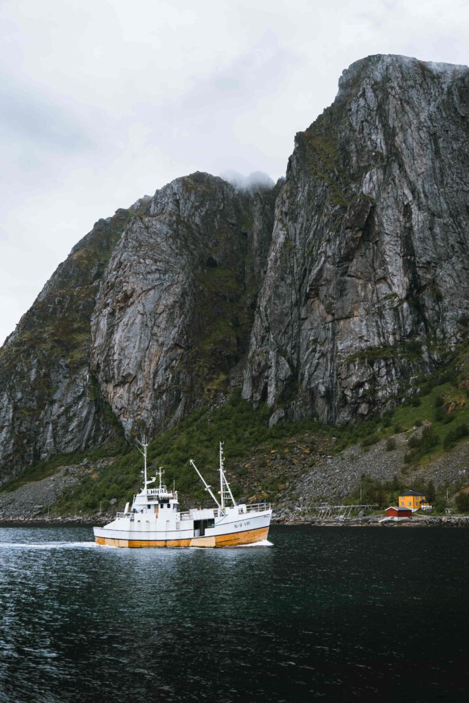 Ett vitt fiskefartyg med gula detaljer seglar i en fjord omgiven av höga berg.