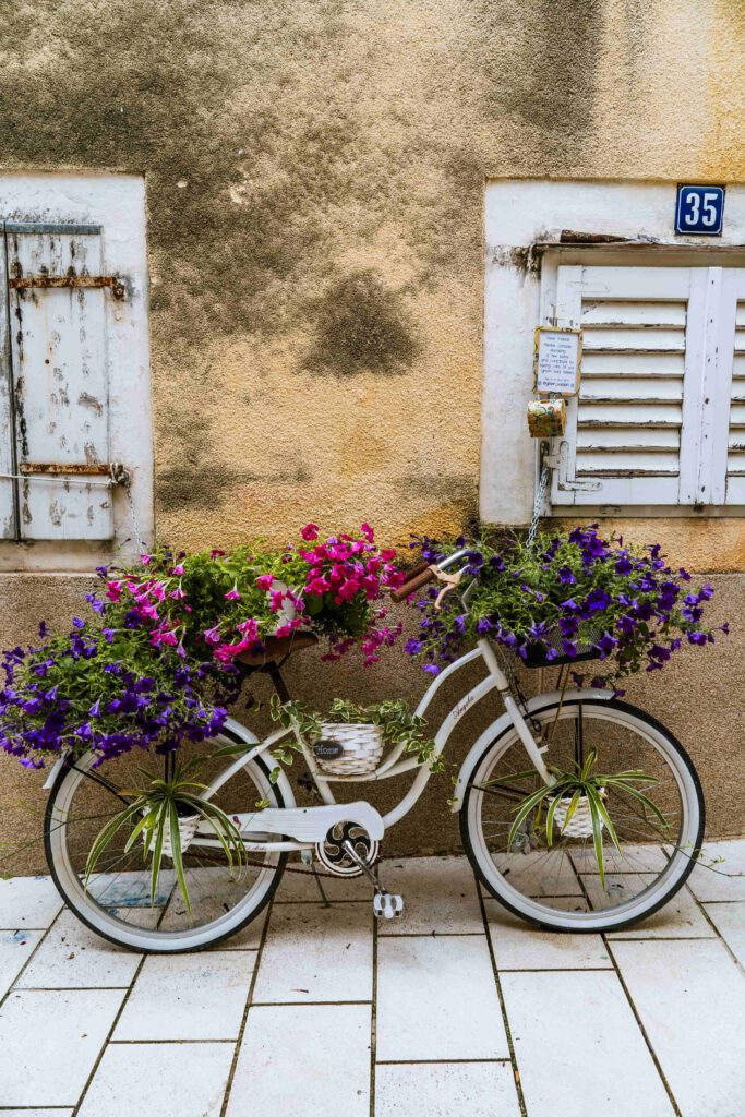 En vit cykel dekorerad med blommor står lutad mot en vägg med slitna fönsterluckor.