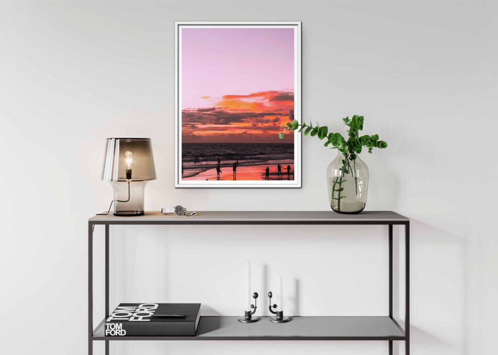 Poster som visar Solnedgång vid havet med rosa och orange himmel, människor som promenerar på stranden, speglade i den våta sanden.