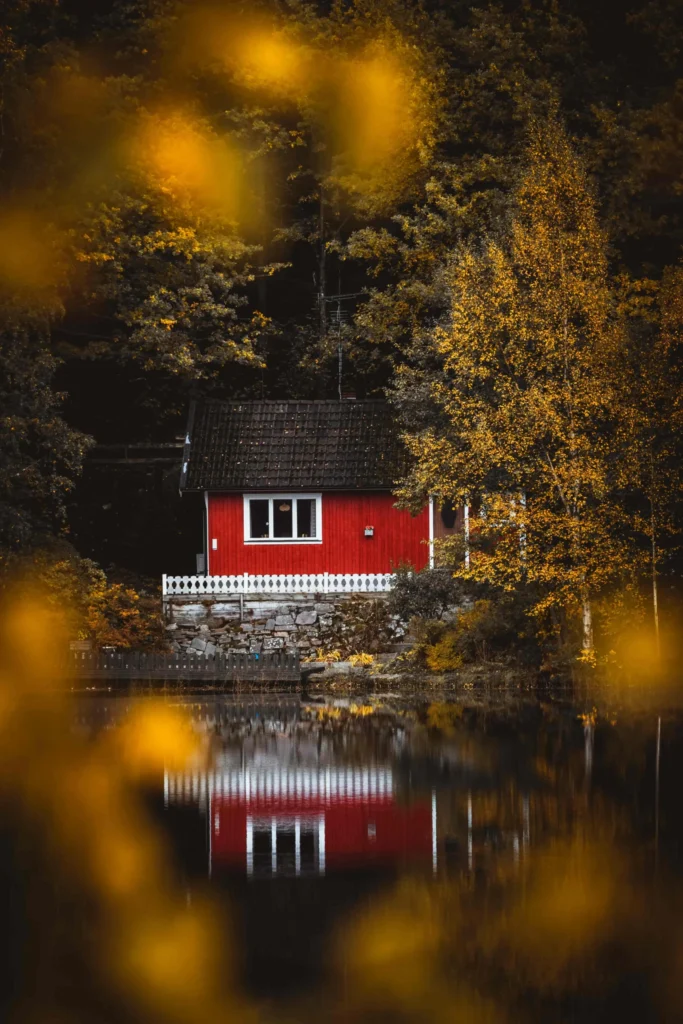 En röd stuga vid en sjö omgiven av höstens gyllene träd, speglad i det lugna vattnet, med oskarpa guldfärgade löv i förgrunden.