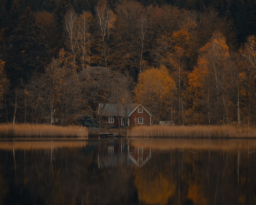 En röd stuga med vita knutar ligger vid en sjö omgiven av höga träd i höstfärger.