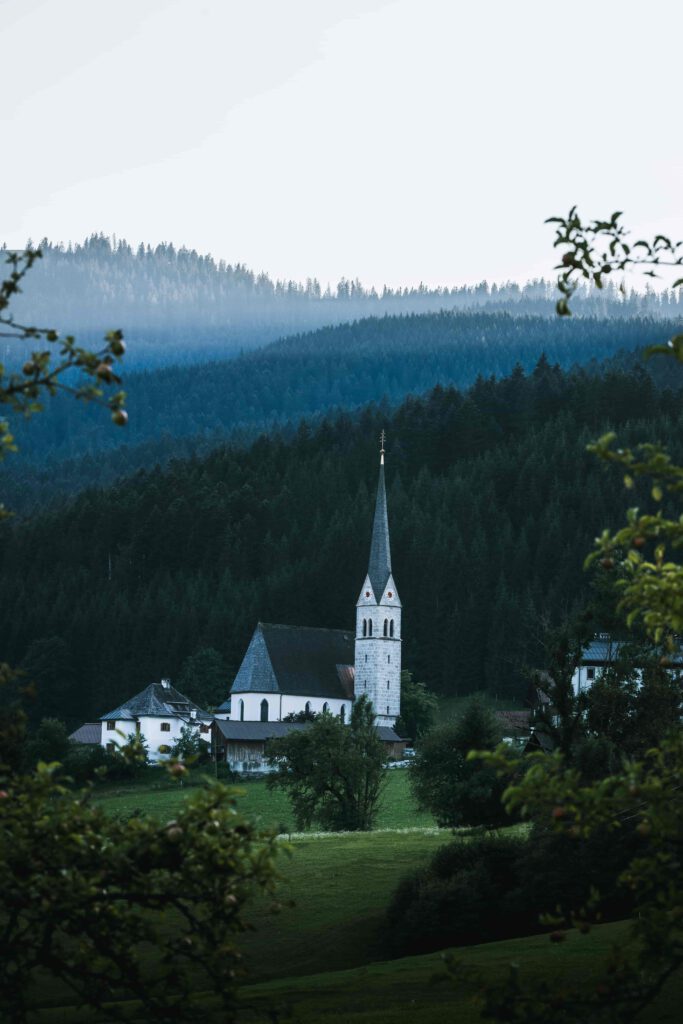 En vit kyrka med ett högt torn ligger i en grön dal omgiven av skog.