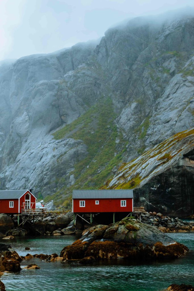 Två röda fiskebodar på pålar vid en norsk fjord med ett berg i bakgrunden.