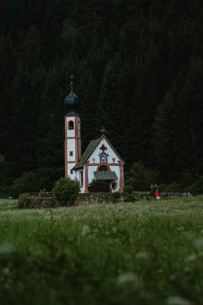 En liten vit kyrka med ett grönt tak och ett spetsigt torn står på en kulle omgiven av gröna fält och en tät skog.